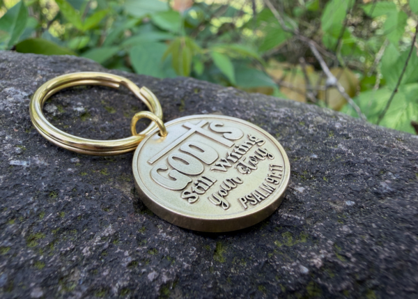 Gold keychain on rock engraved with God is still writing your story. Psalm 91:11 amidst greenery.