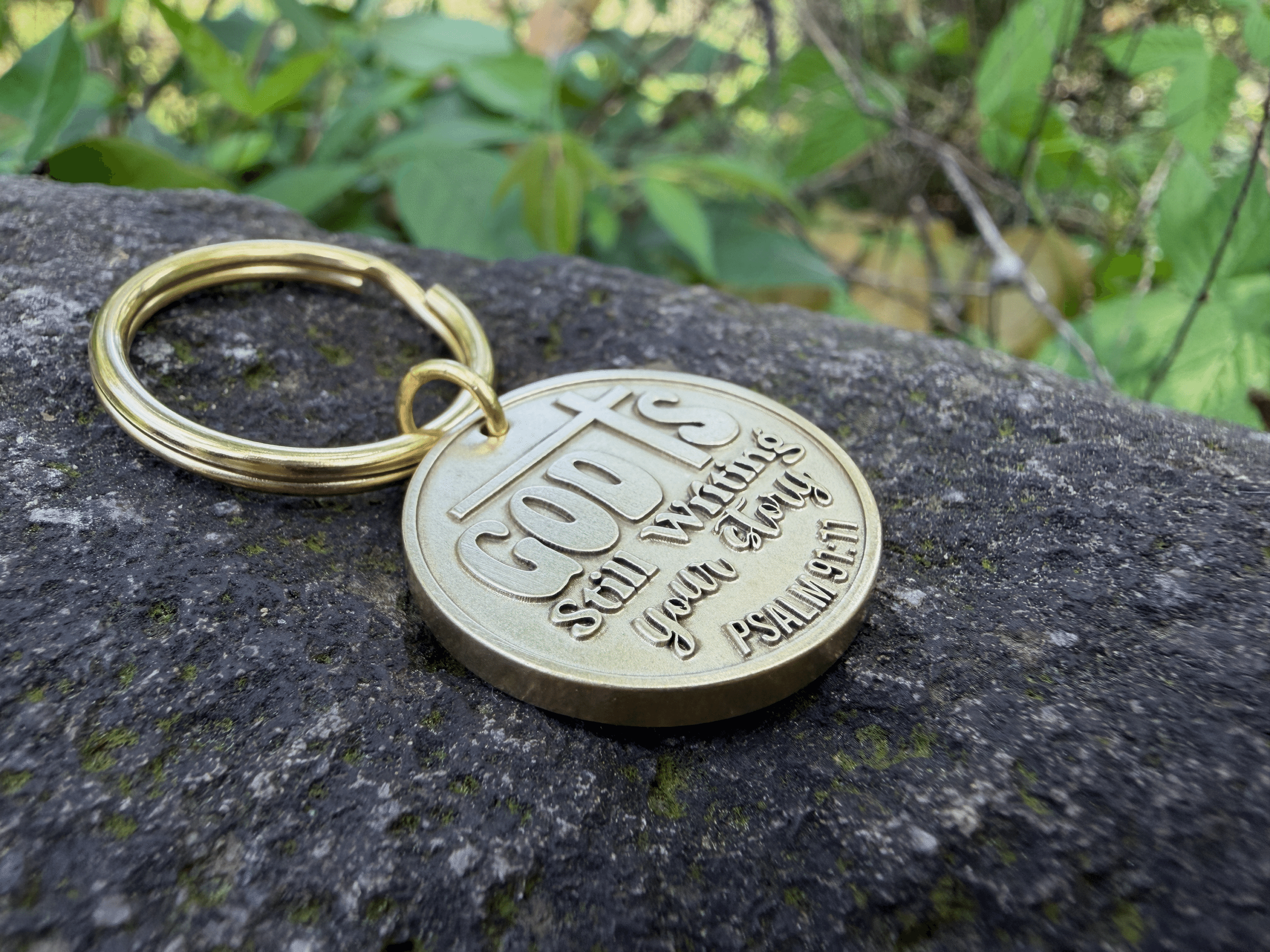 Gold keychain on rock engraved with God is still writing your story. Psalm 91:11 amidst greenery.