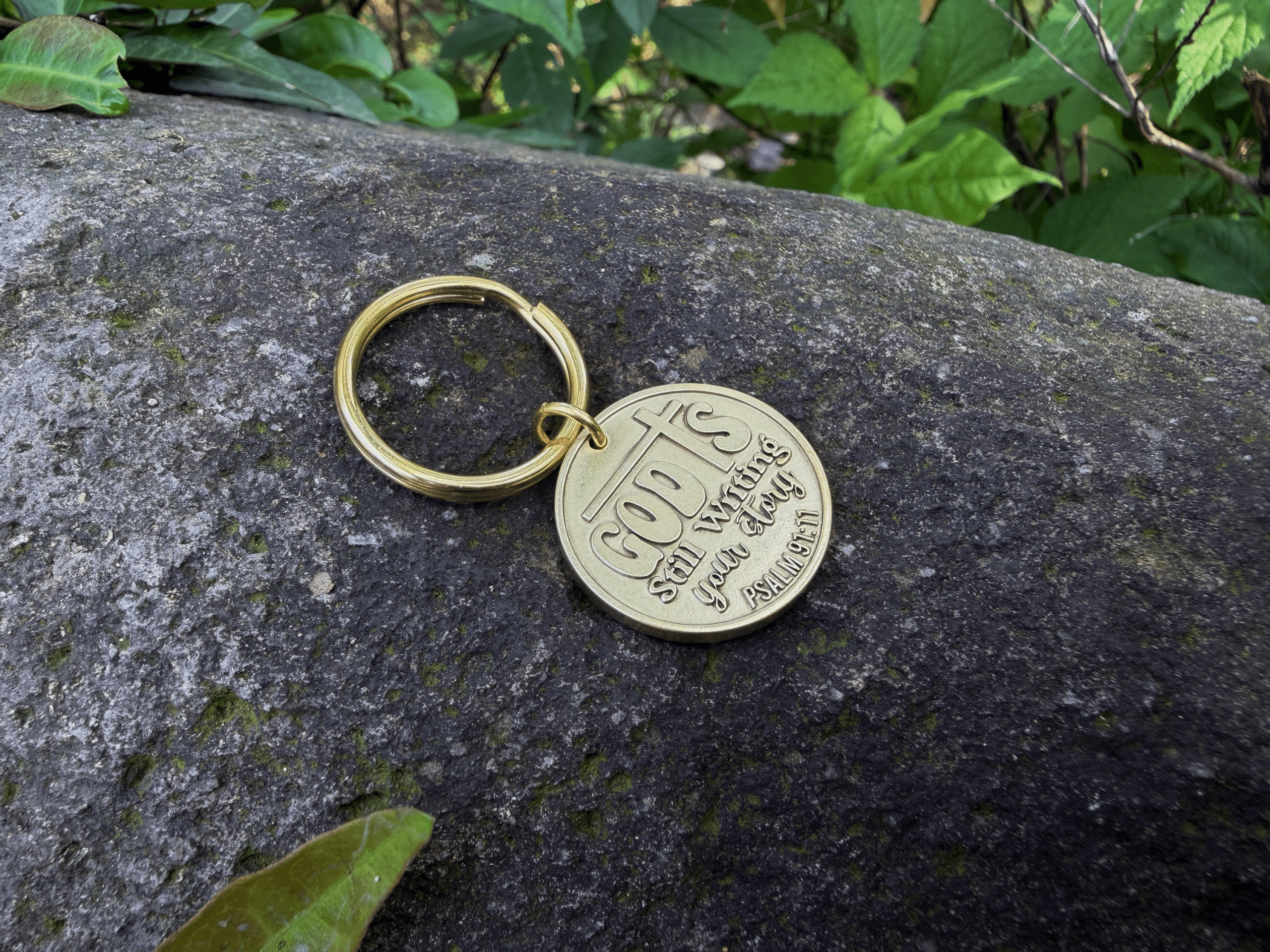 Gold keychain on a rock engraved with God's Still Writing Your Story. Psalm 139:16. in a green natural setting.