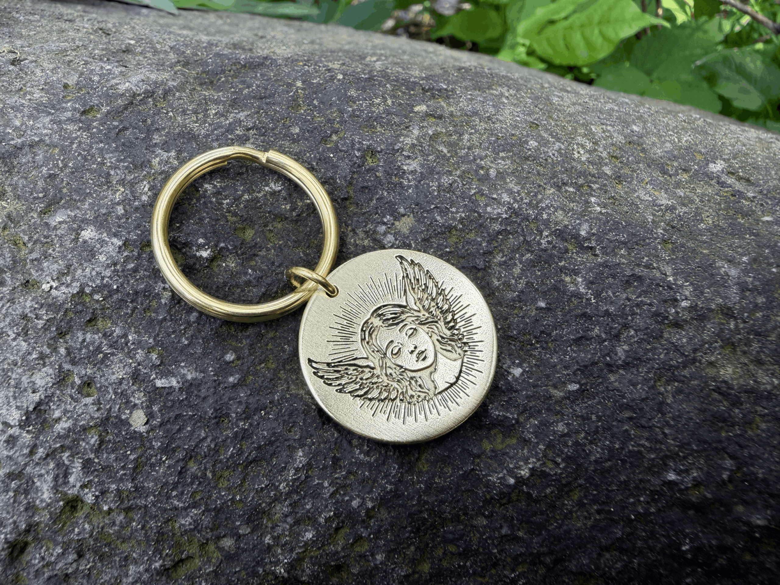 Gold keychain with engraved angel face and wings on a stone surface.