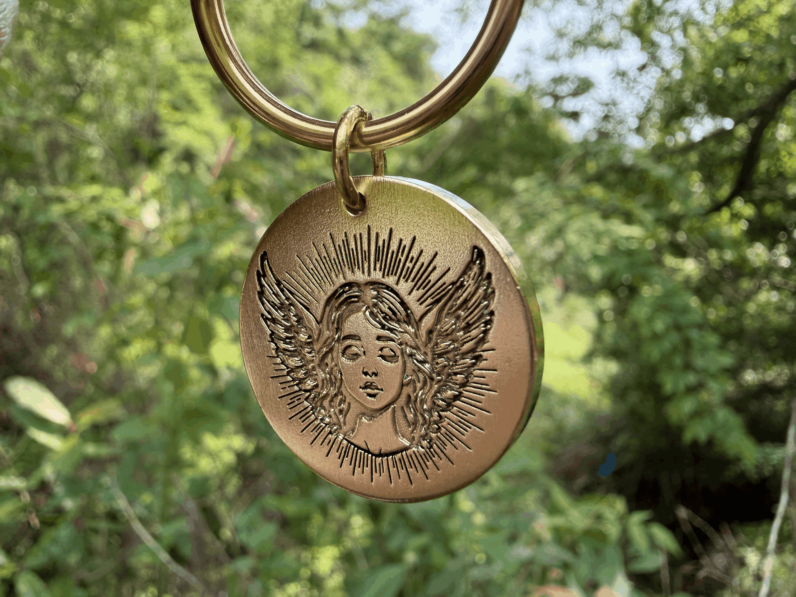 Golden keychain with angel engraving, wings spread, sunlight glinting in a lush green forest backdrop.