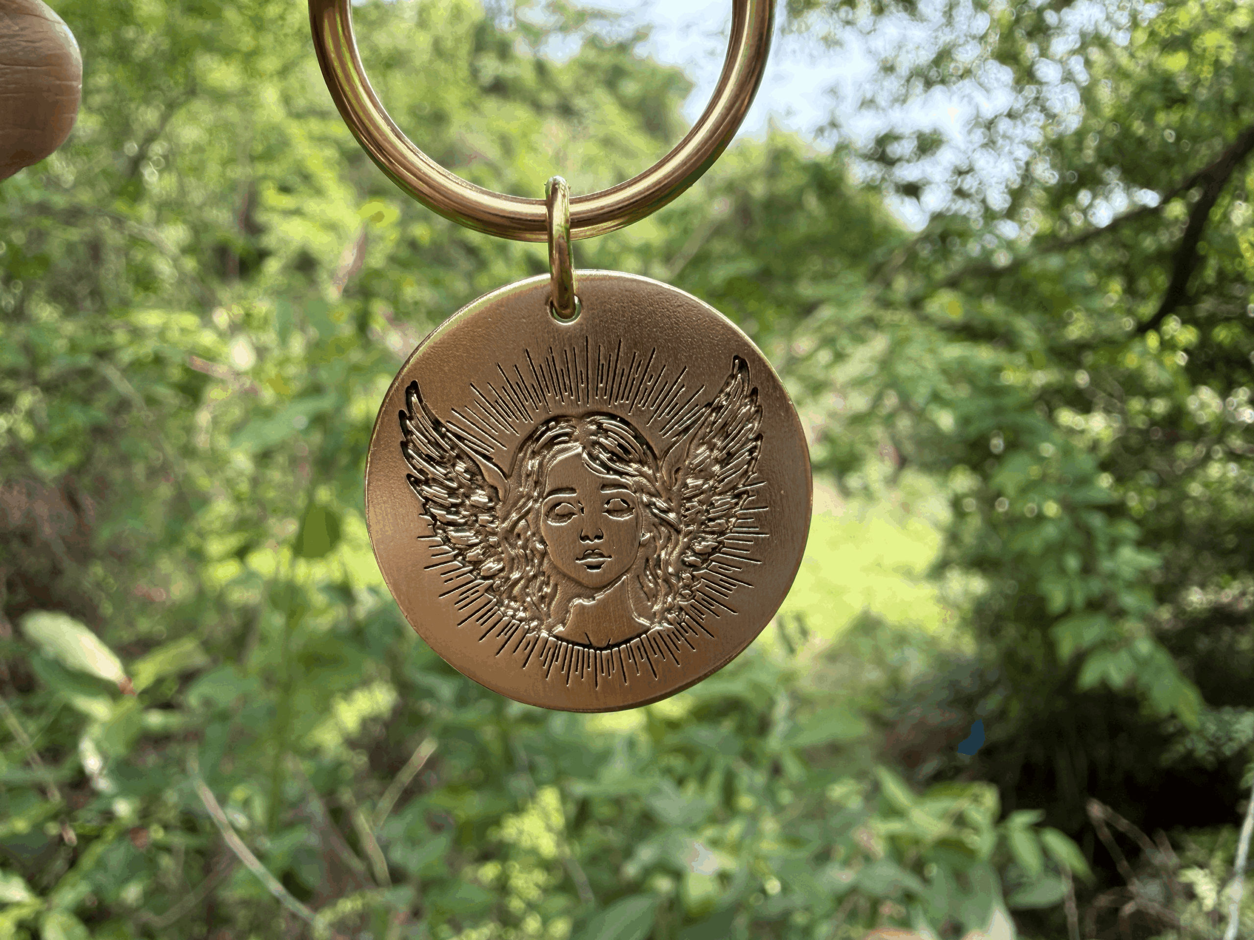 Golden medallion with engraved angelic face and wings, surrounded by sun rays, hanging outdoors against a lush green background.