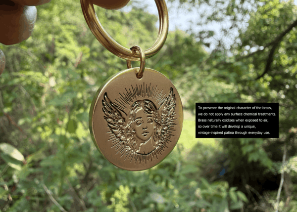 Brass keychain with angel engraving, hanging outdoors, highlighting natural, untreated patina development over time.