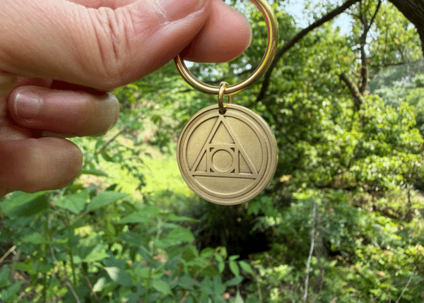 Hand holding a brass keychain engraved with the AA Recovery symbol, set against a lush, green forest backdrop.