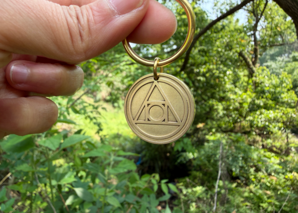 Hand holding a brass keychain engraved with the AA Recovery symbol, set against a lush, green forest backdrop.