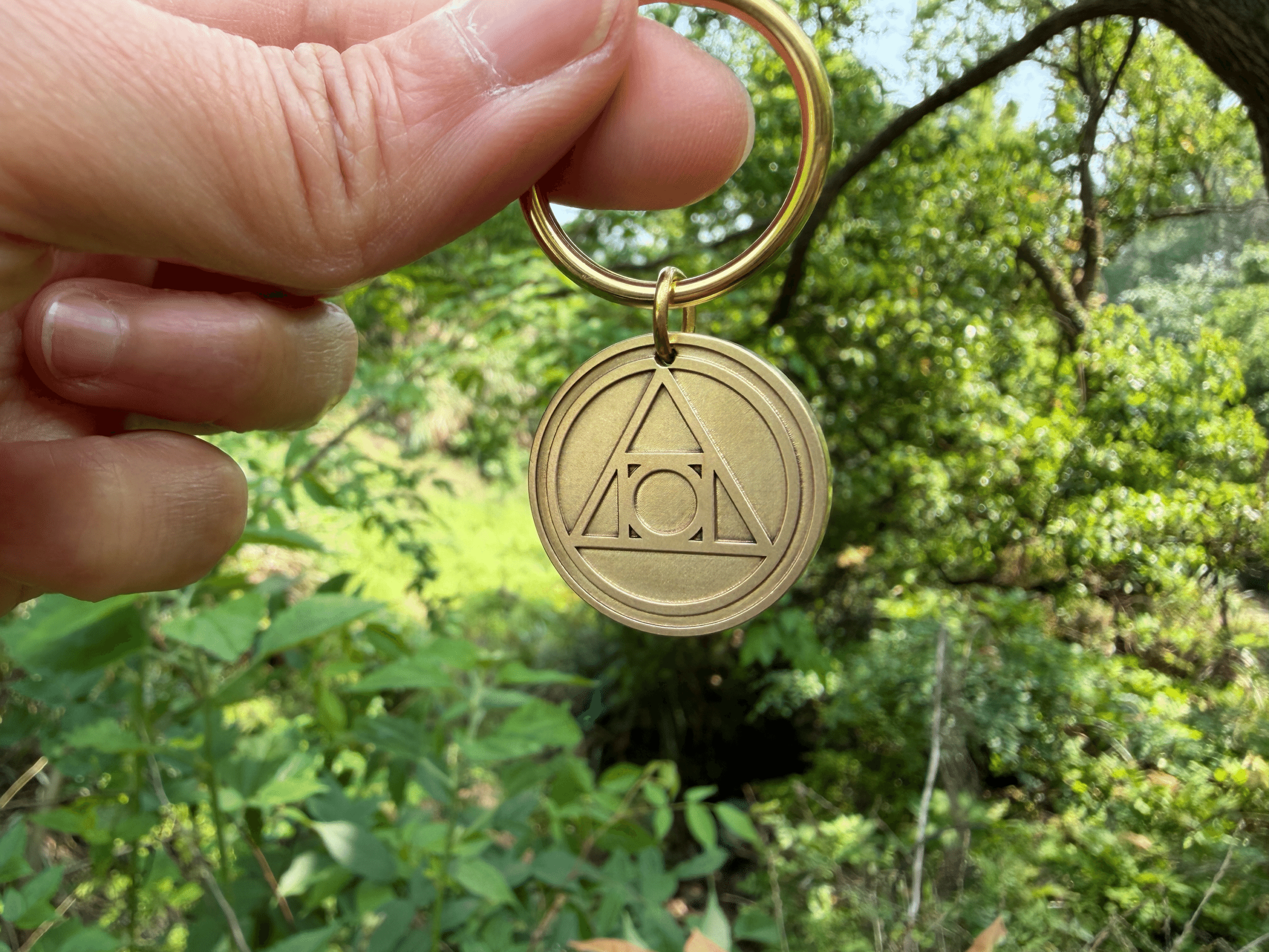 Hand holding a brass keychain engraved with the AA Recovery symbol, set against a lush, green forest backdrop.