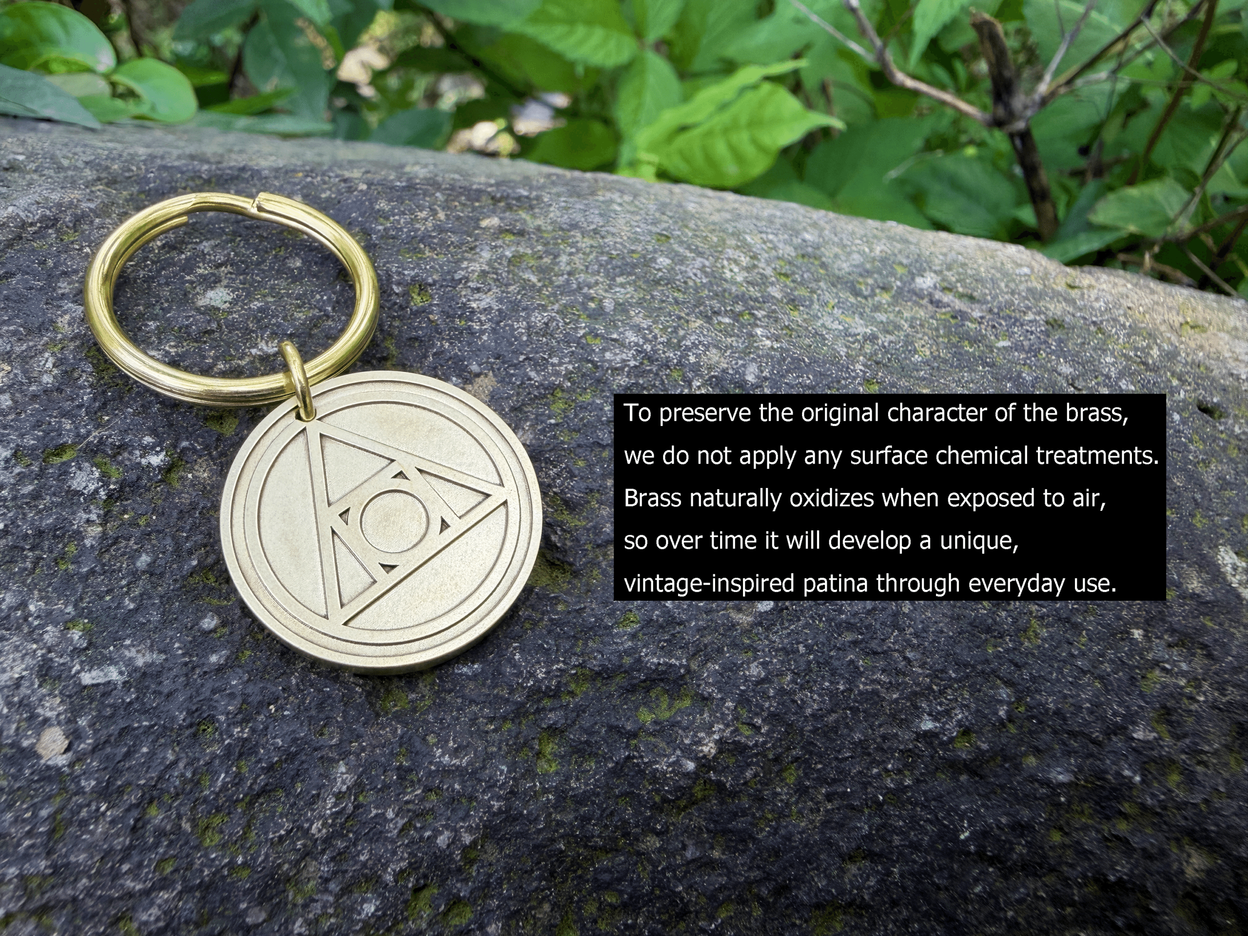 Golden brass keychain with AA Recovery engraved symbol on rock, surrounded by green leaves.