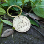 Gold keychain with a circle and triangle engraving, symbolizing the AA Recovery, resting on green leaves.