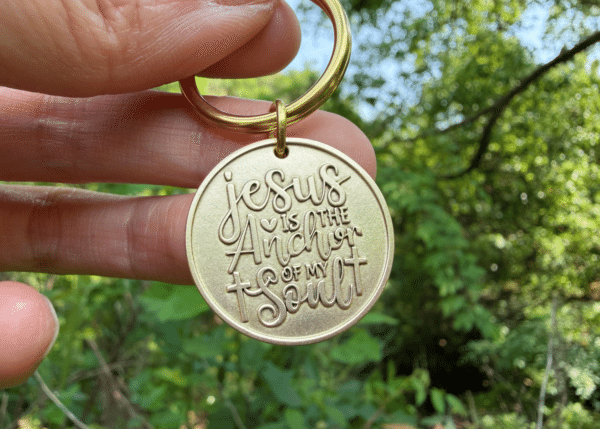 Gold keychain engraved with Jesus is the Anchor of My Soul held outdoors with greenery in the background.