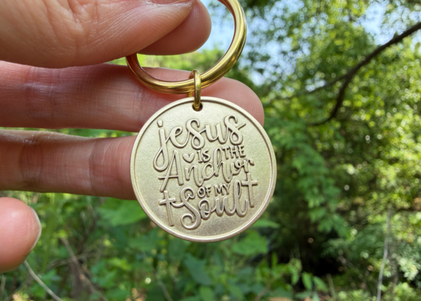 Gold keychain engraved with Jesus is the Anchor of My Soul held outdoors with greenery in the background.