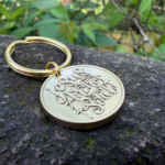 Gold keychain on stone with engraving Jesus is the Anchor of my Soul, set against lush green foliage background.