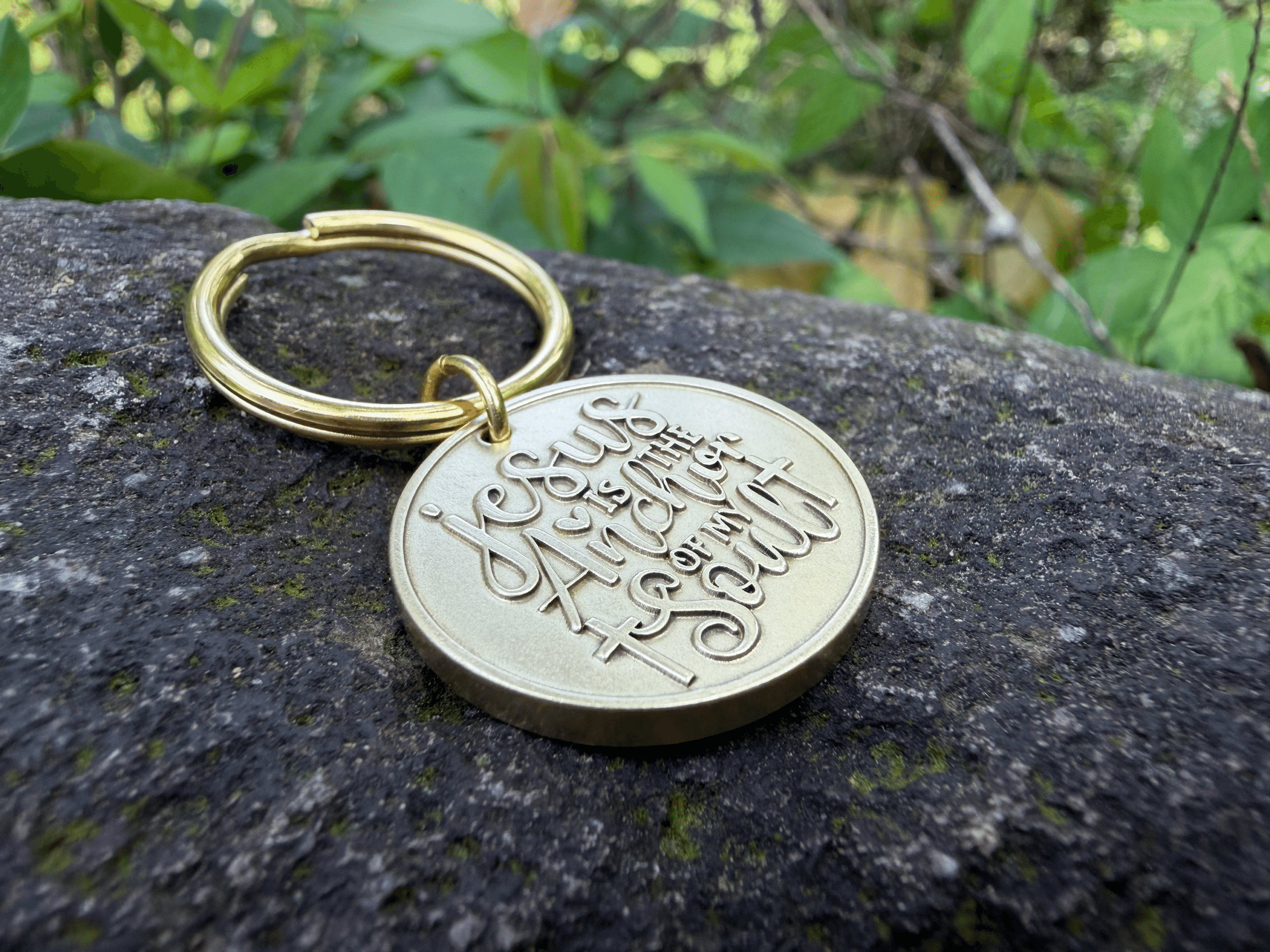Gold keychain on stone with engraving Jesus is the Anchor of my Soul, set against lush green foliage background.
