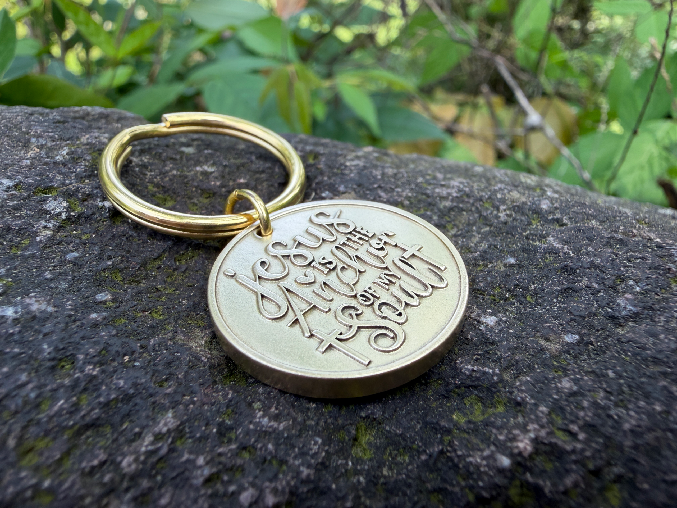 Gold keychain on stone with engraving Jesus is the Anchor of my Soul, set against lush green foliage background.