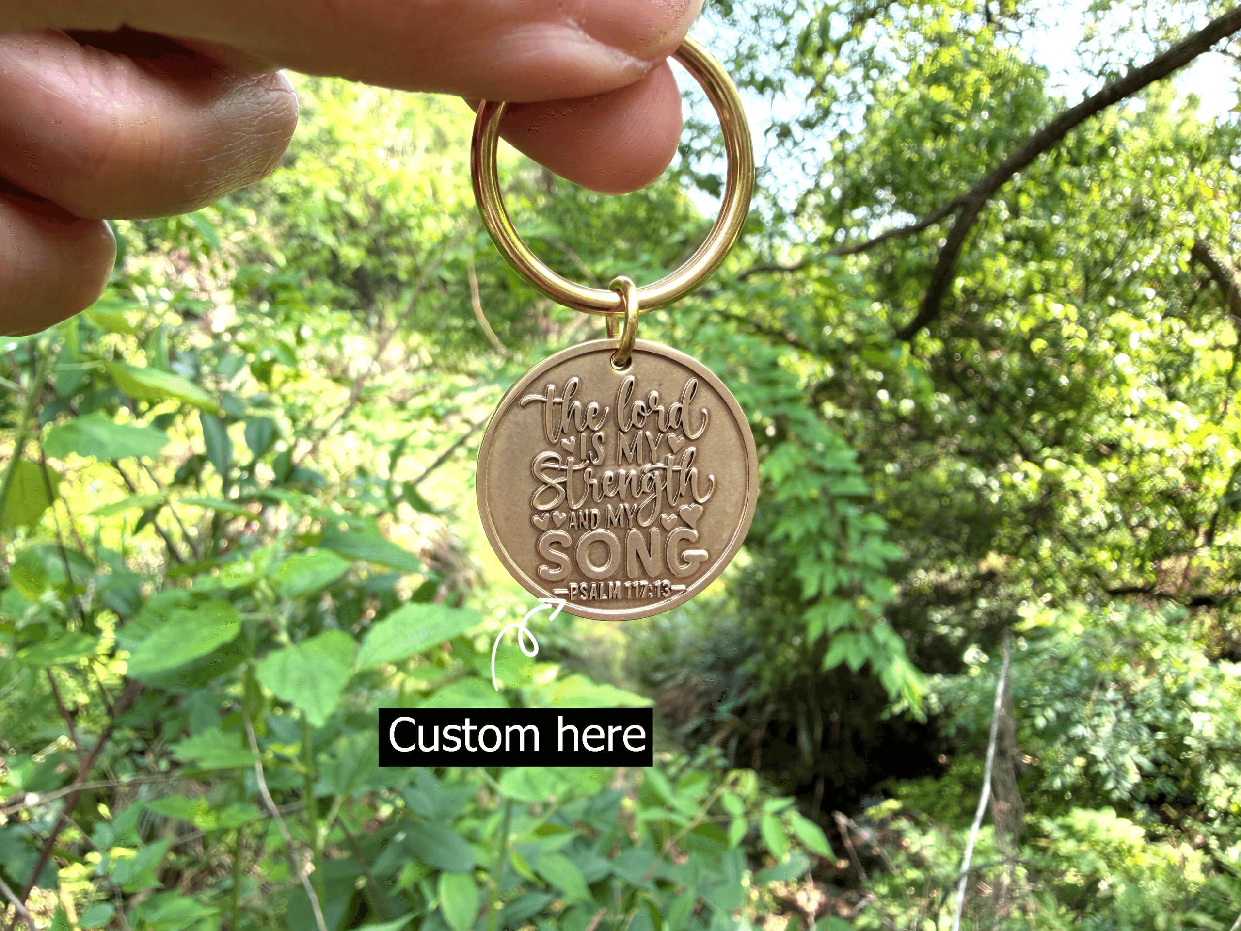 Gold keychain with engraving The Lord is my strength and my song - Psalm 117:13 held against a leafy background.