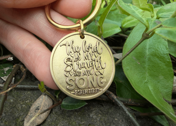 Keychain engraved with The Lord is my strength and my song, Psalm 117:13, held outdoors near leaves.