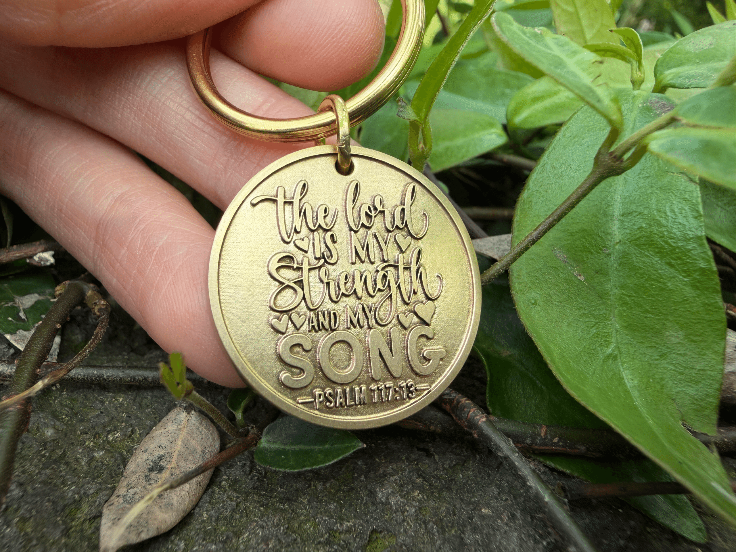 Keychain engraved with The Lord is my strength and my song, Psalm 117:13, held outdoors near leaves.