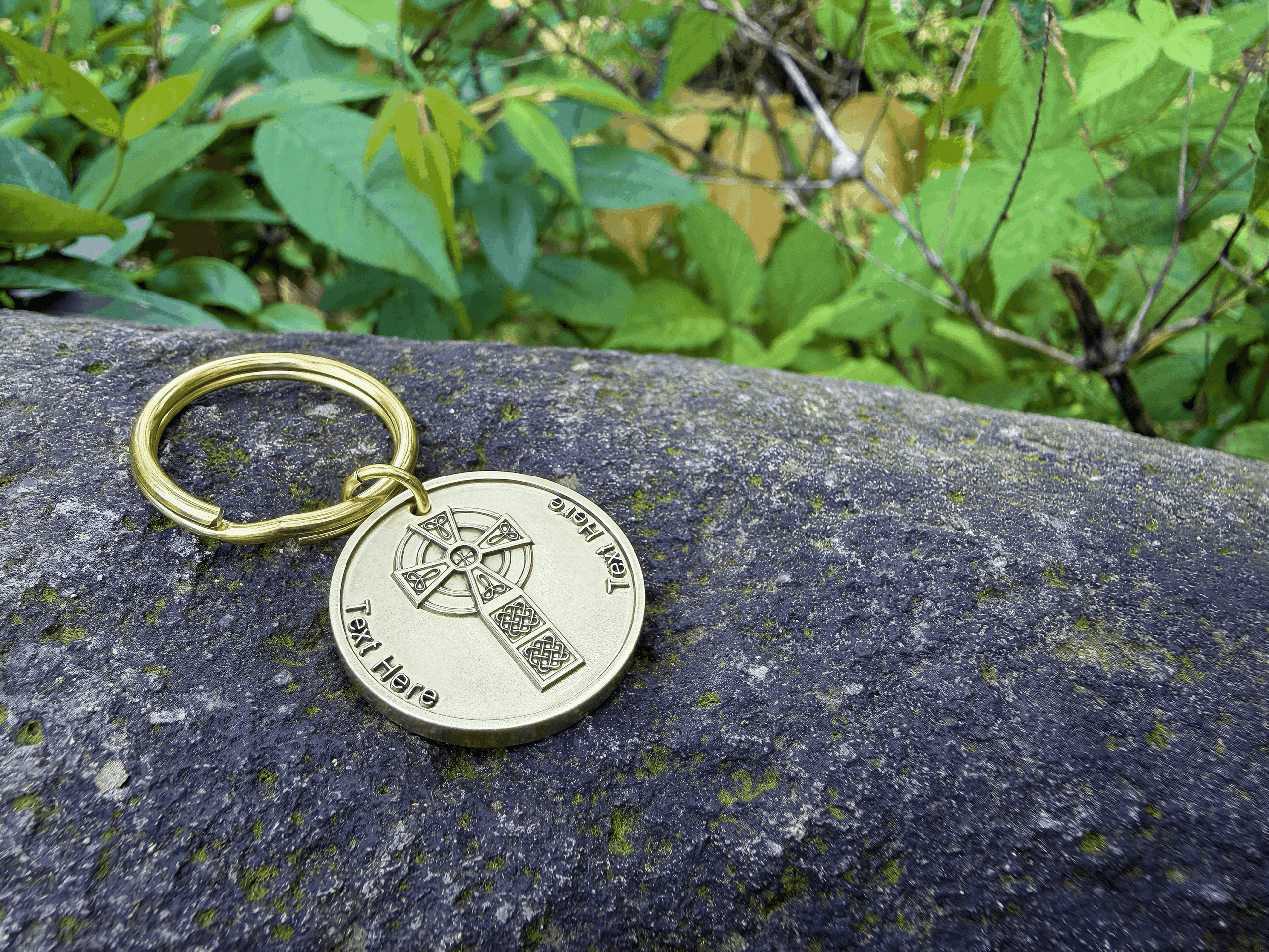 Gold keychain with engraved Celtic cross resting on a mossy rock, surrounded by green foliage in nature.