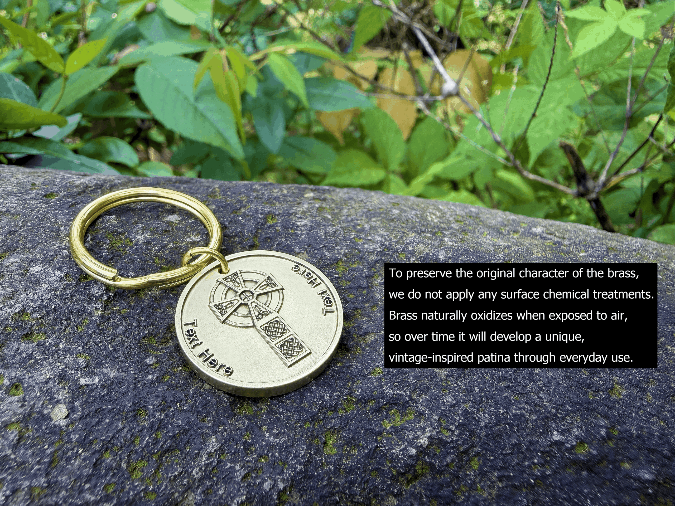 Brass Celtic Cross keychain with engraved cross design on stone, surrounded by green foliage. Text describes natural oxidation and patina.