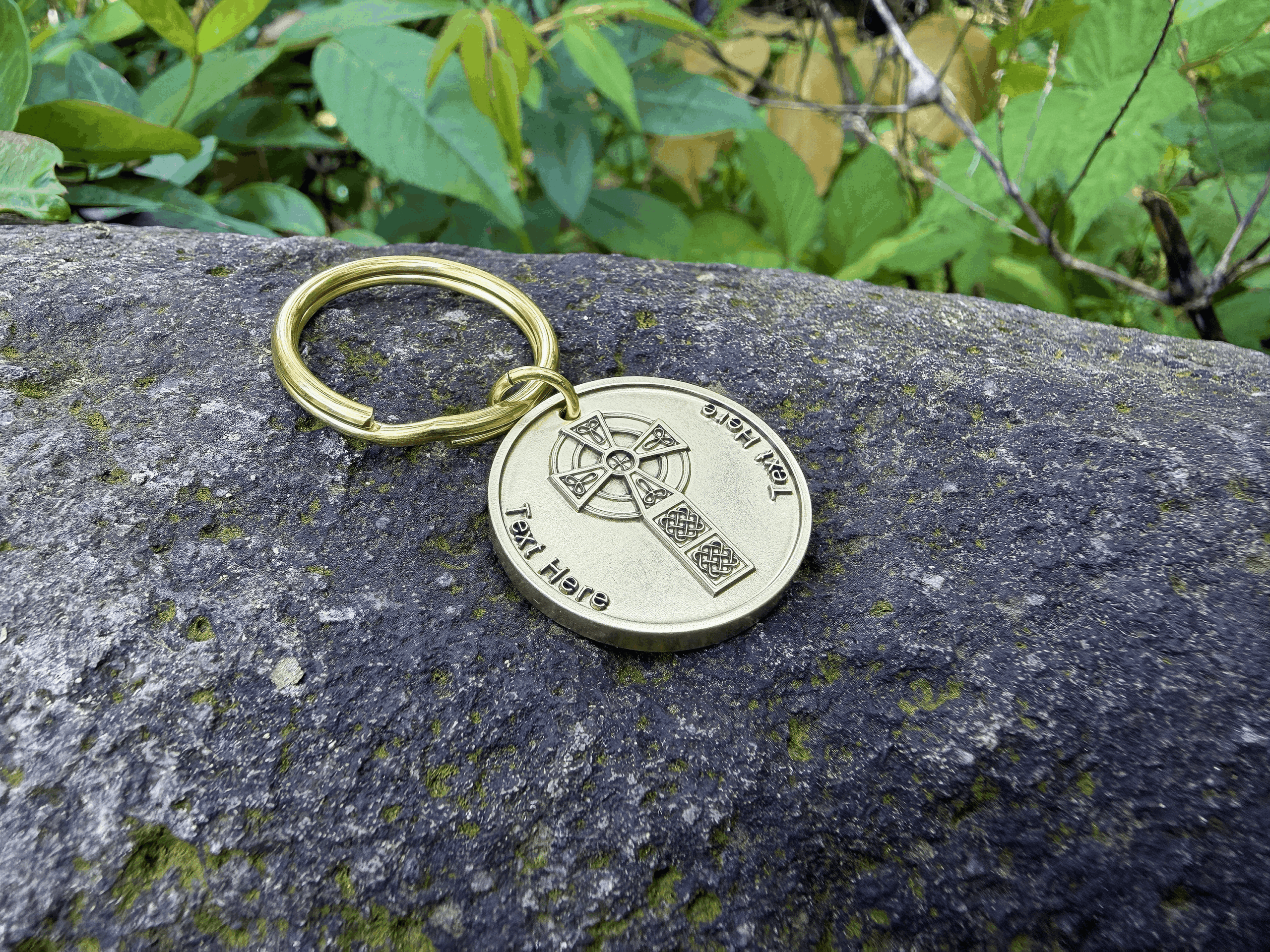 Gold keychain with Celtic Cross design and customizable text, resting on a mossy rock in a lush garden setting.