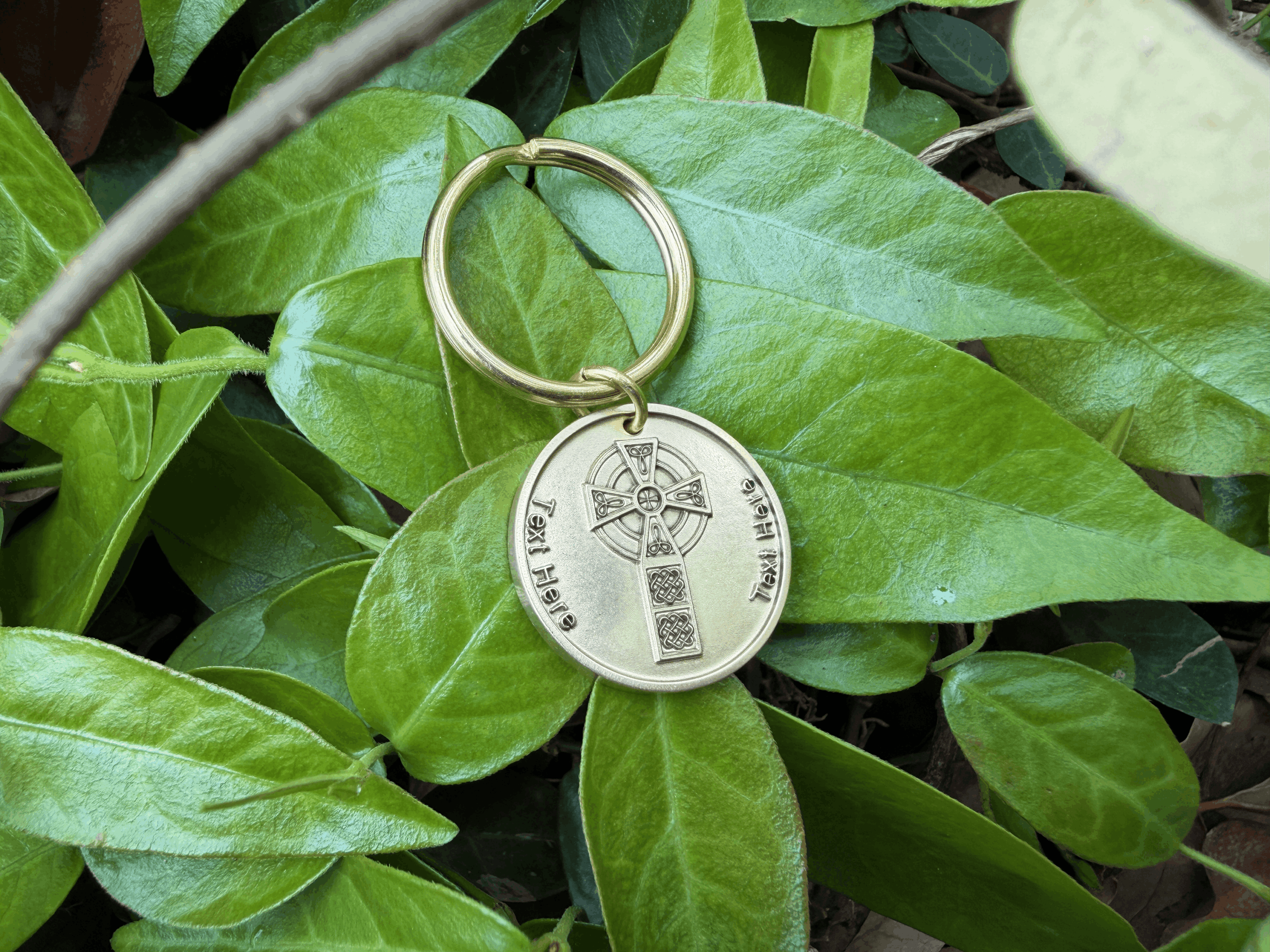 Gold Celtic Cross keychain with intricate cross design resting on vibrant green leaves.