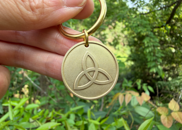 Hand holding a gold keychain with engraved Celtic triquetra symbol, lush green background.