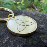 Gold keychain with Celtic Triquetra engraving placed on a mossy stone surface outdoors.
