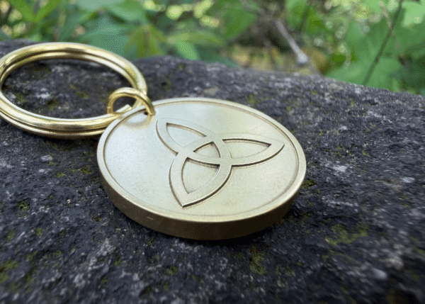 Gold keychain with Celtic Triquetra engraving placed on a mossy stone surface outdoors.