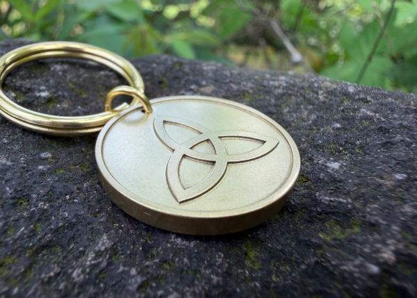 Gold keychain with Celtic Triquetra engraving placed on a mossy stone surface outdoors.