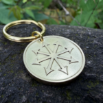 Gold metal keychain with chaos star engraving resting on a rock with greenery in the background.