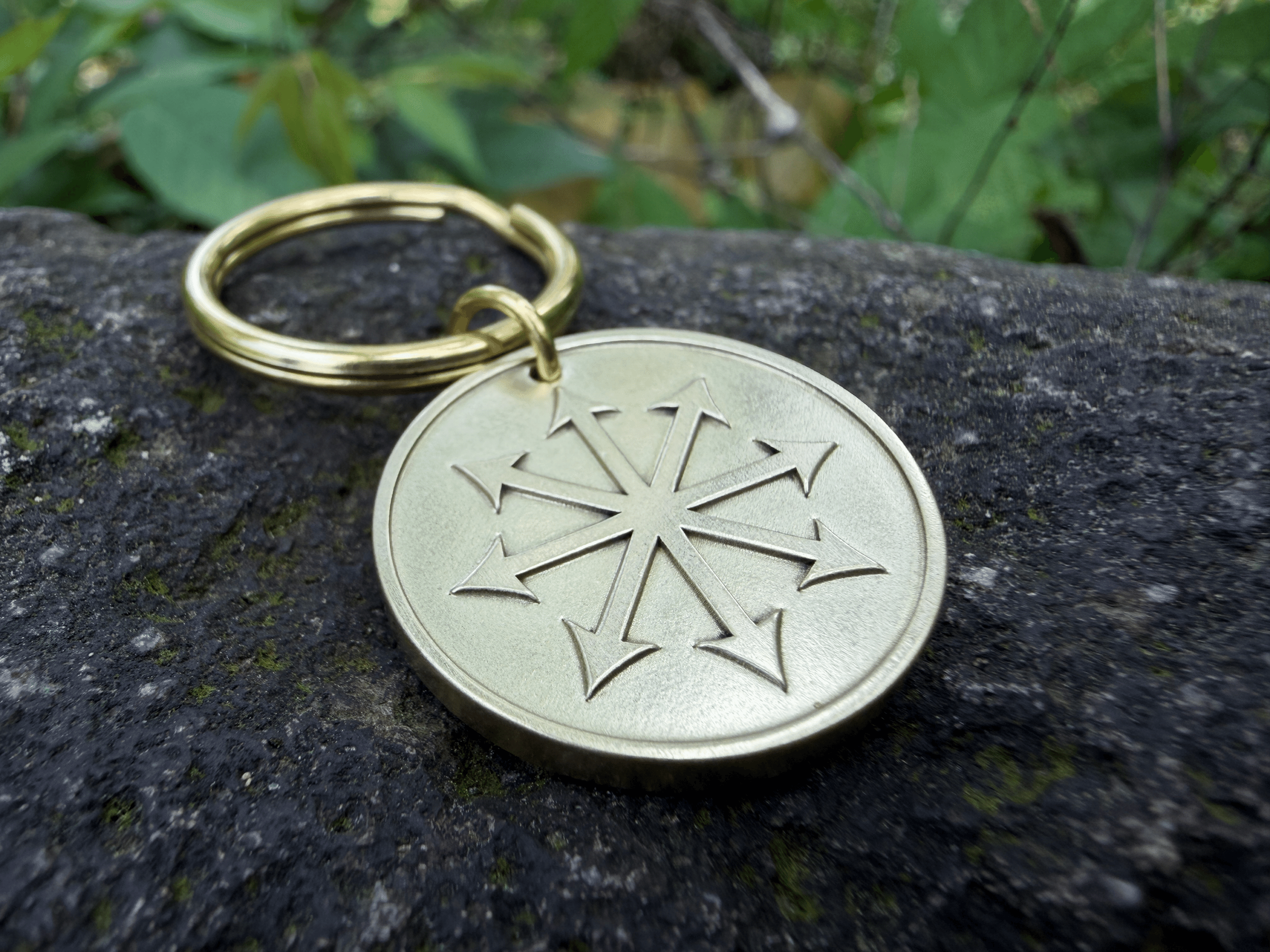 Gold metal keychain with chaos star engraving resting on a rock with greenery in the background.