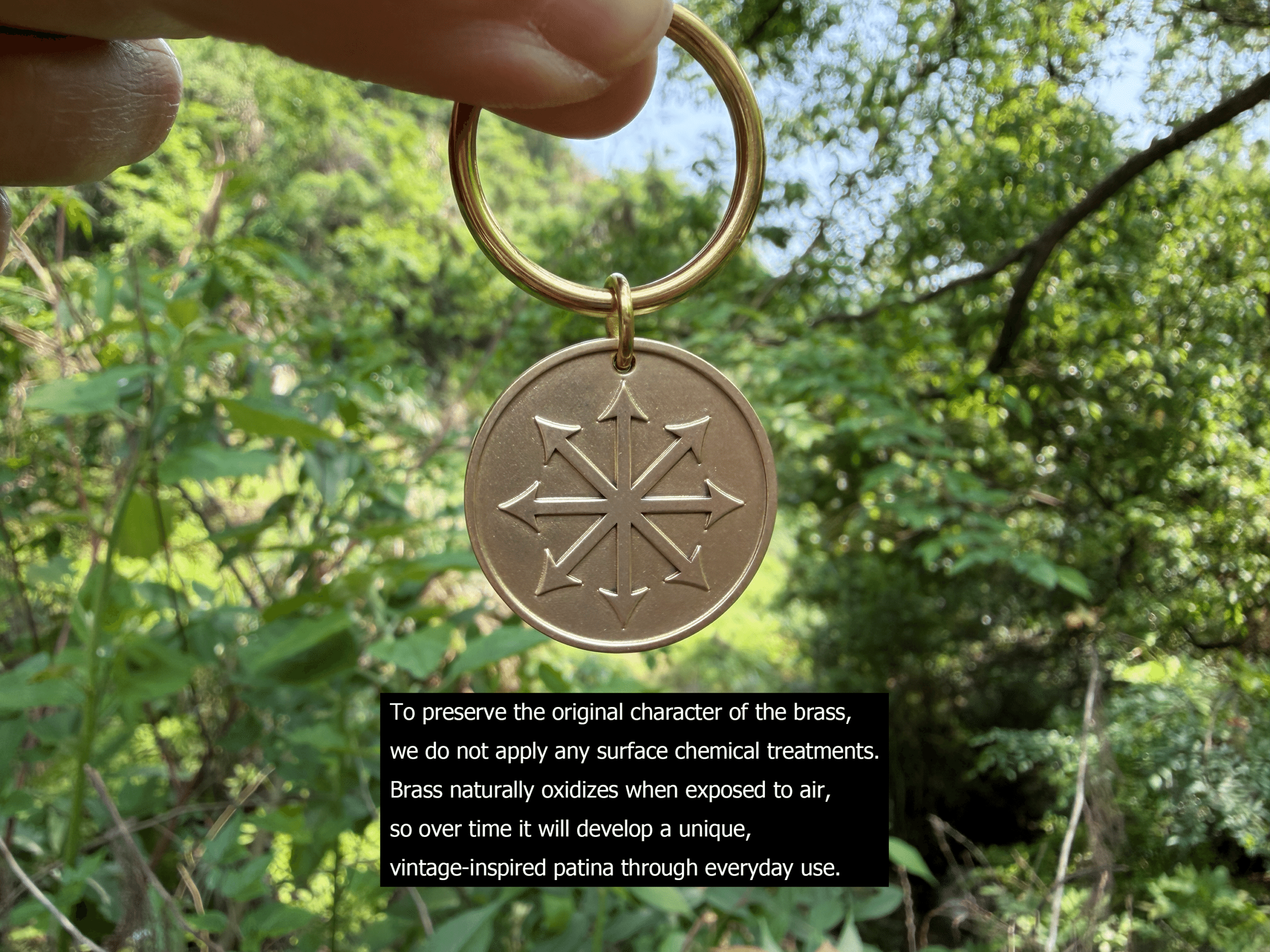 Brass keychain with engraved compass rose, held against a lush green forest background. Text about brass patina.