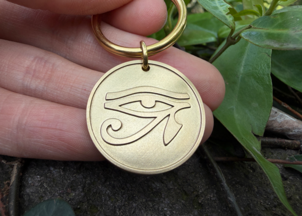Golden keychain engraved with Eye of Horus held between fingers amid green foliage.