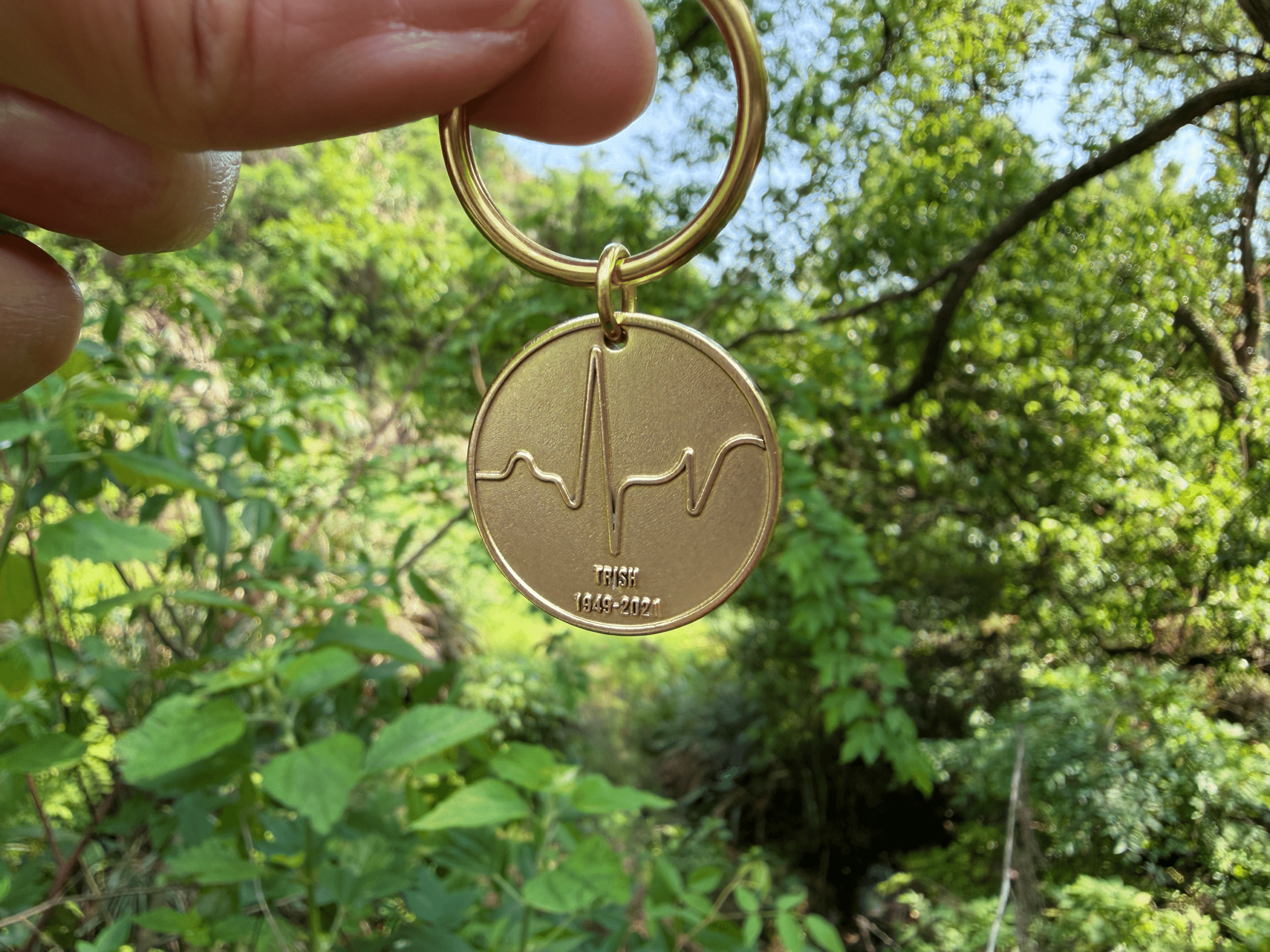 Gold keychain with heartbeat engraving, memorial text TRISH 1949-2021, held against a green leafy background.