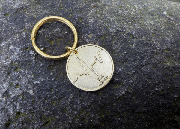 Gold keychain with wave engraving and text IRISH 1949-2021 on a stone surface.