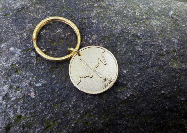 Gold keychain with wave engraving and text IRISH 1949-2021 on a stone surface.