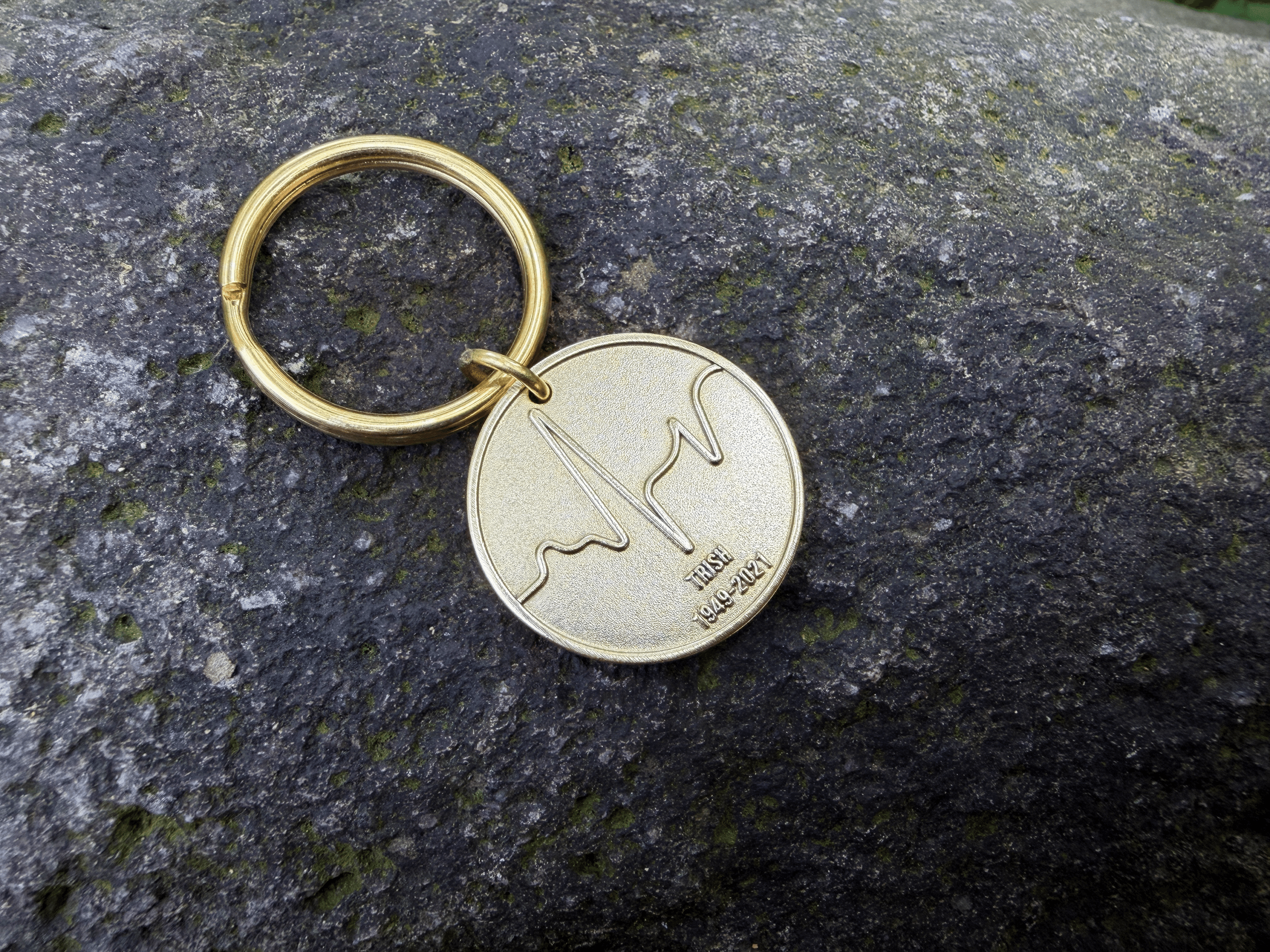 Gold keychain with wave engraving and text IRISH 1949-2021 on a stone surface.