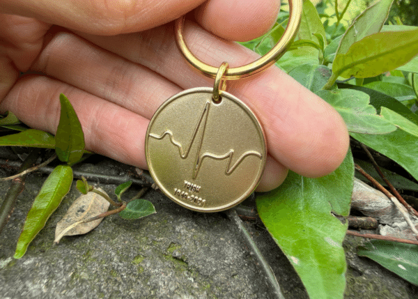 Gold keychain with engraved heartbeat line and the text TRISH 1949-2021, held by fingers above green leaves.
