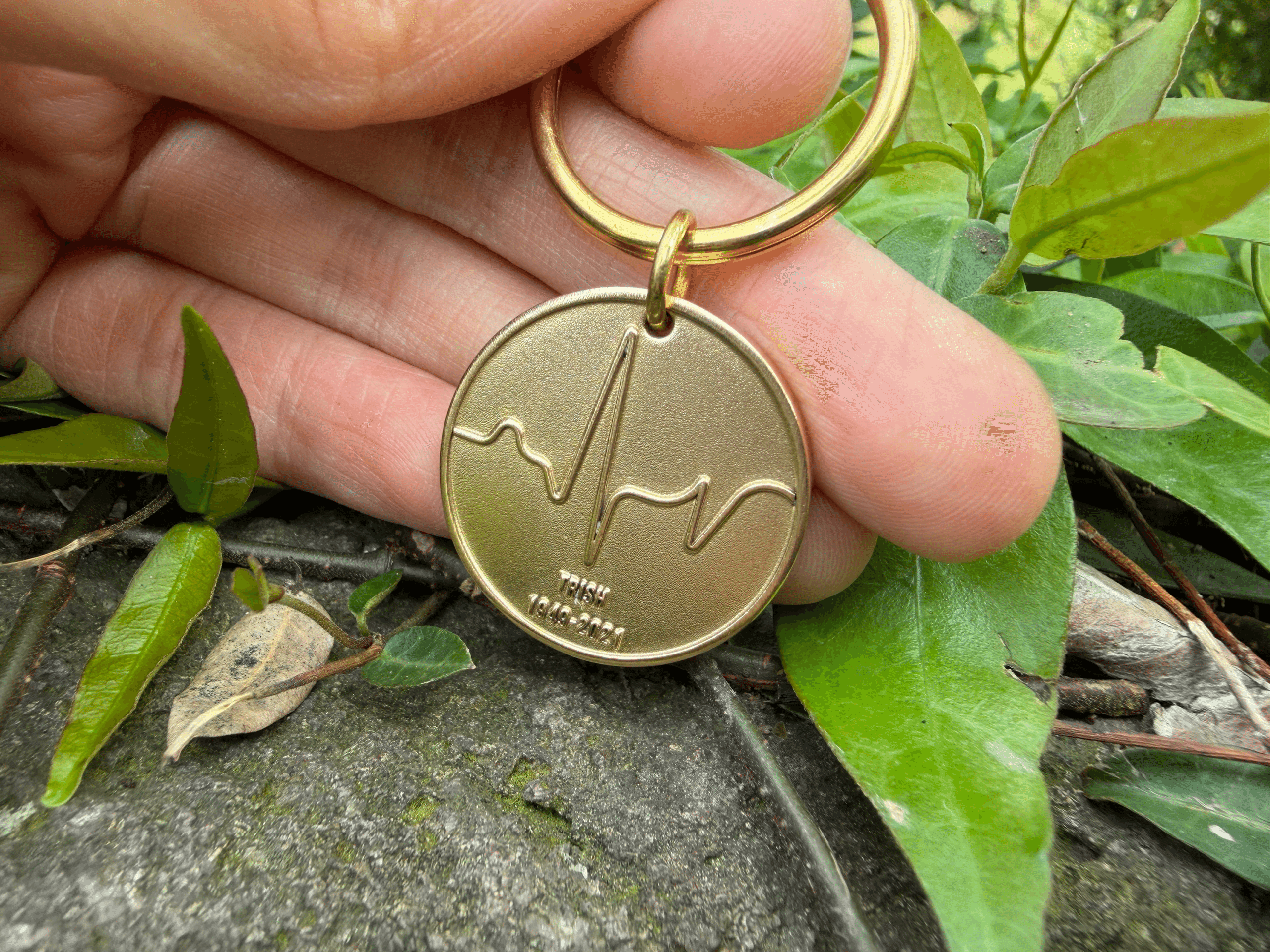 Gold keychain with engraved heartbeat line and the text TRISH 1949-2021, held by fingers above green leaves.