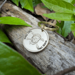 Keychain with skull and crossbones engraving, Text Here, placed on wood surrounded by green leaves.