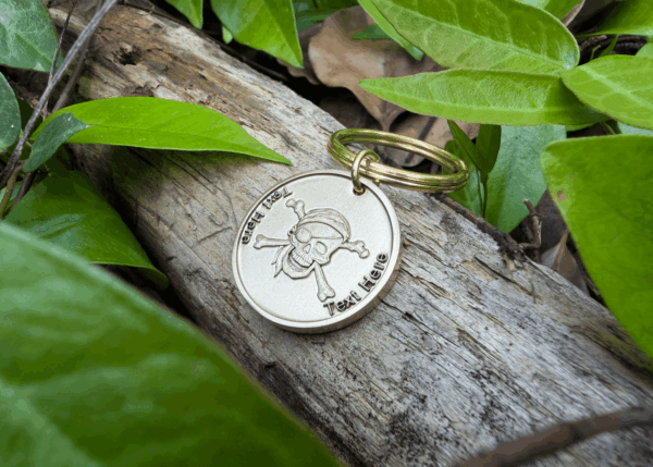 Keychain with skull and crossbones engraving, Text Here, placed on wood surrounded by green leaves.