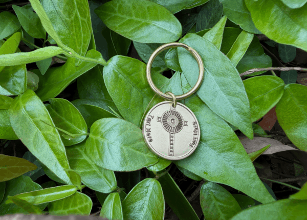 Personalized Monstrance Cross Keychain with circular medallion on lush green leaves background, featuring engraved geometric design.