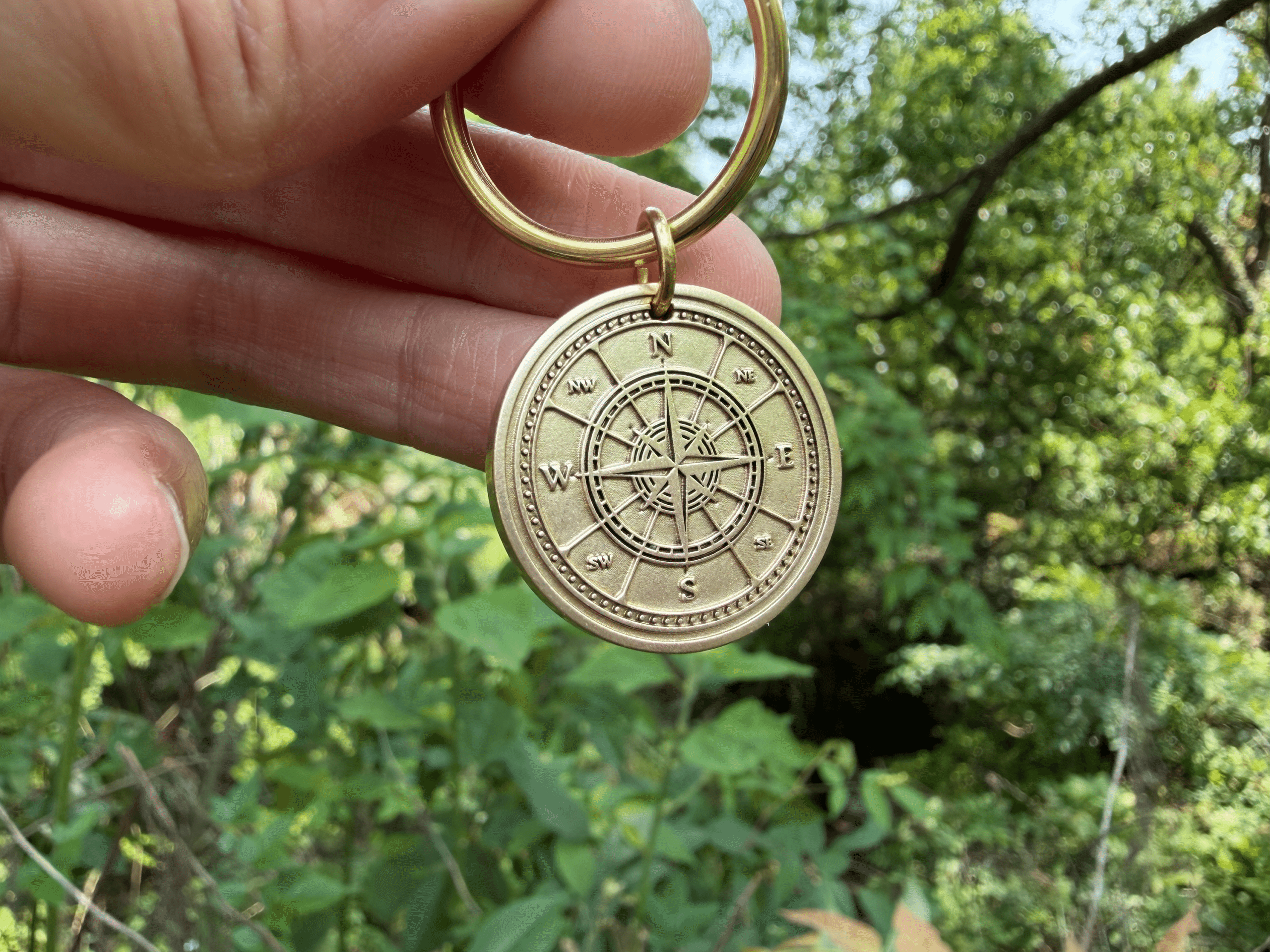 Hand holding a compass keychain with cardinal directions engraving in a lush, green outdoor setting.