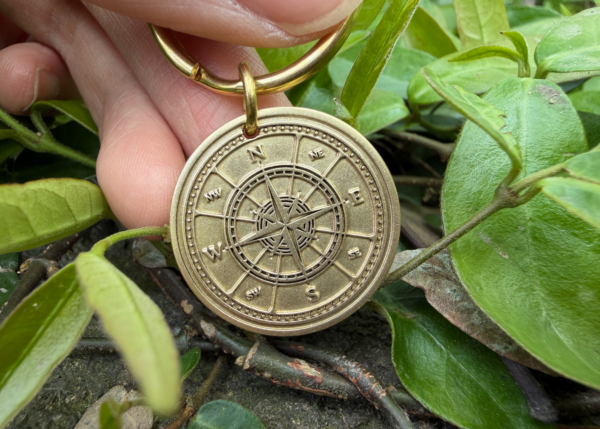 Hand holds brass compass keychain with engraved directional markings, surrounded by green leaves.