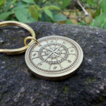 Gold keychain with engraved compass design on a rock surface, surrounded by green leaves.