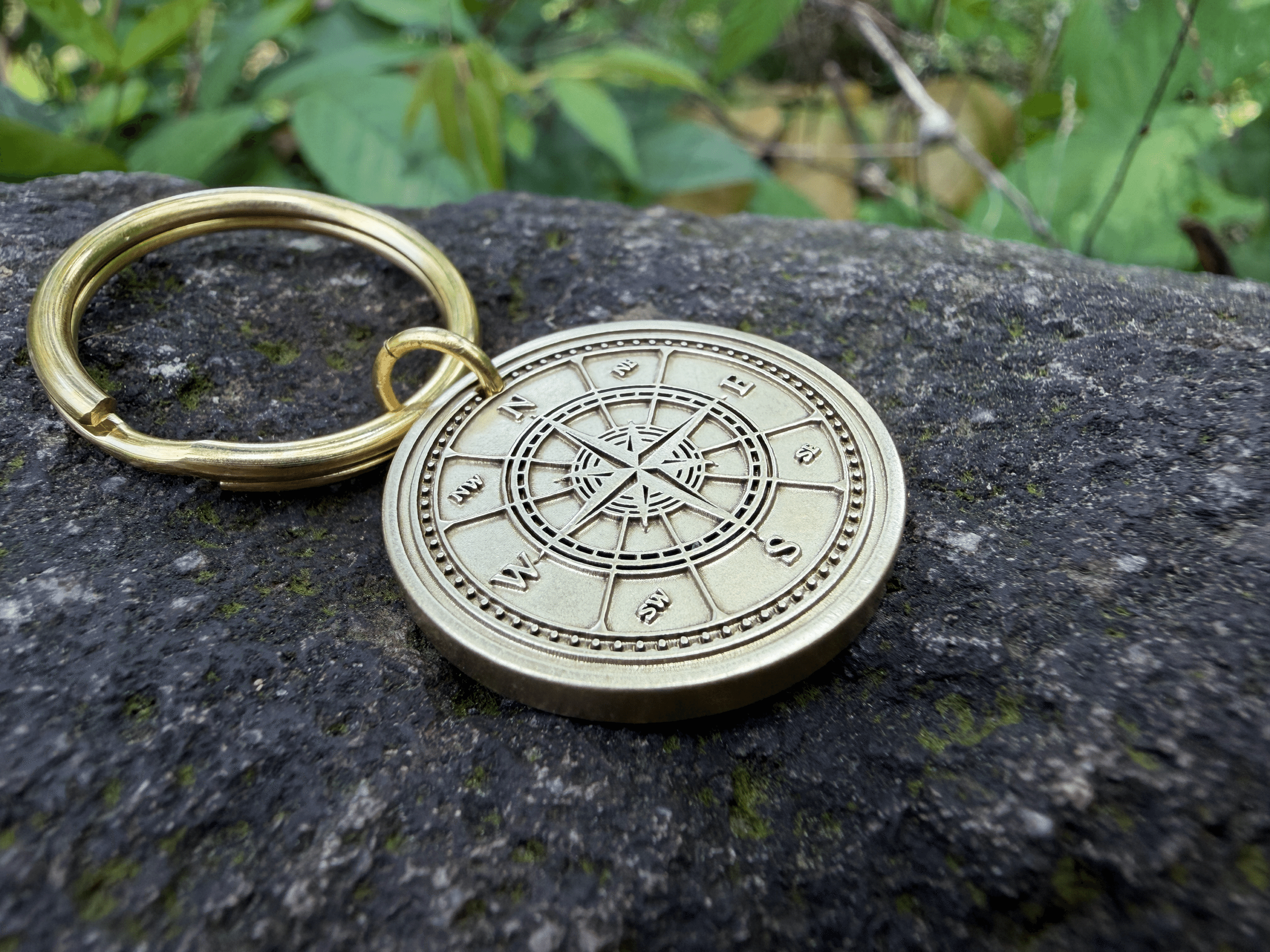 Gold keychain with engraved compass design on a rock surface, surrounded by green leaves.