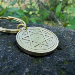 Gold keychain with Star of David engraving resting on a stone, surrounded by greenery.