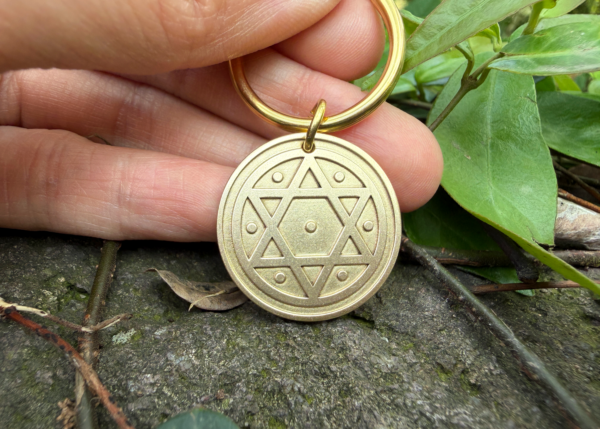 Bronze keychain with hexagram and star engravings, held by hand, placed on a stone with green leaves.