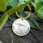 Gold keychain with tree engraving and date 03.21.2025, resting on green leaves and stone background.