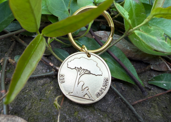 Gold keychain with tree engraving and date 03.21.2025, resting on green leaves and stone background.