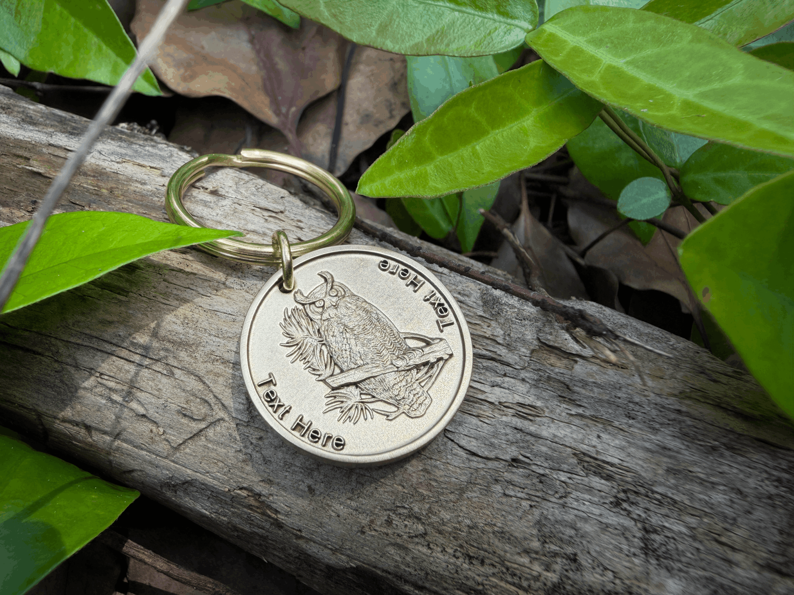 Keychain on wooden log with engraved owl design and customizable text, surrounded by green leaves.
